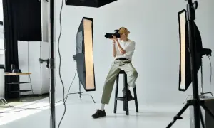 Młody fotograf z blond włosami, siedząc na stołku, robi zdjęcie aparatem. W studiu widać lampy i sprzęt. Zastanawiasz się, ile zarabia fotograf?
