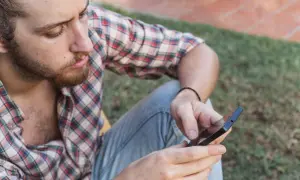 Mężczyzna w kratę szybko sprawdza powiadomienia na telefonie, siedząc na trawie.