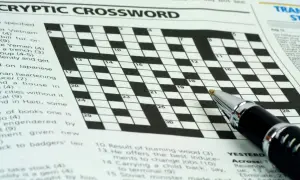 Zbliżenie na krzyżówkę z długopisem, widoczne nagłówki "CRYPTIC CROSSWORD" i fragmenty zadań.