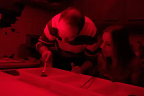 W ciemnym pokoju, przy czerwonym świetle, mężczyzna i dziewczyna pracują przy wywoływaniu zdjęć.