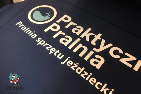 Druk termotransferowy na granatowej odzieży: logo pralni "Praktyczna Pralnia" i napis "Pralnia sprzętu jeździeckiego".