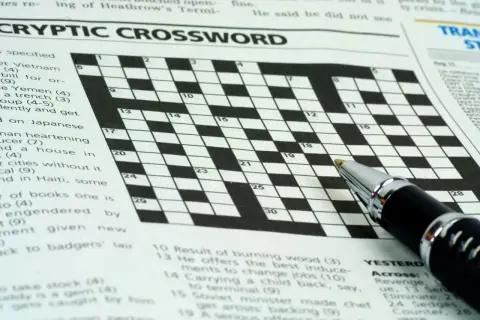 Zbliżenie na krzyżówkę z długopisem, widoczne nagłówki "CRYPTIC CROSSWORD" i fragmenty zadań.