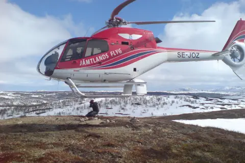 Czerwony helikopter Jämtlands Flyg SE-JOZ wylądował na zaśnieżonym wzgórzu. Fotograf robi zdjęcie, uchwycony przez efekt rolling shutter.