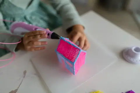 Dziecko tworzy różowy domek z niebieskim dachem za pomocą długopisu 3D. Powstaje piękny, trójwymiarowy ornament.