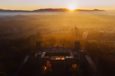 Zamek otoczony mgłą, skąpany w ciepłych promieniach **golden hour**. Słońce wschodzi nad wzgórzami, malując niebo na złoto.