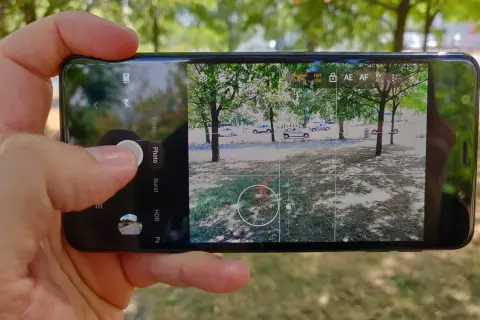 Dłoń trzyma smartfon z otwartą aplikacją do robienia zdjęć. Widok na drzewa i samochody.