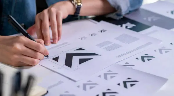 Osoba szkicuje logo wektorowe na papierze, otoczona wieloma wydrukami tego samego projektu.