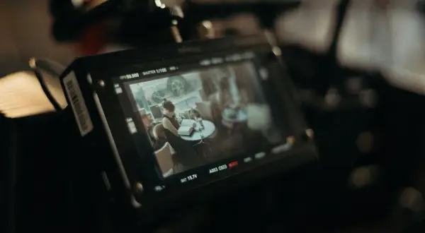Ekran monitora kamery filmowej pokazuje scenę z kobietą czytającą książkę przy stoliku. To ujęcie z planu filmowego.