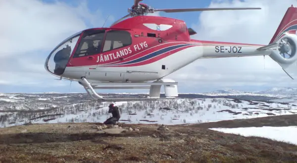 Czerwony helikopter Jämtlands Flyg SE-JOZ wylądował na zaśnieżonym wzgórzu. Fotograf robi zdjęcie, uchwycony przez efekt rolling shutter.