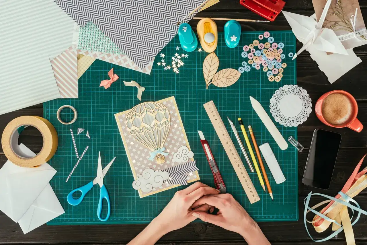 Ręce tworzą kartkę metodą scrapbooking. Widać papiery, nożyczki, taśmę, ozdoby i kawę.