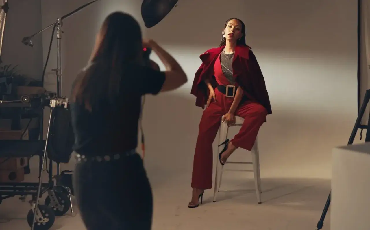 Sesja zdjęciowa w studio. Modelka w czerwonym stroju pozuje na stołku. Fotografka robi zdjęcie. Tak wygląda praca, która pokazuje, ile zarabia fotograf.