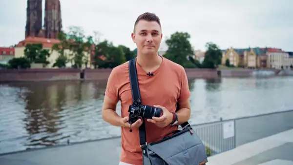 Mężczyzna z aparatem na ramieniu, w tle jasnoniebieskie niebo i rzeka.