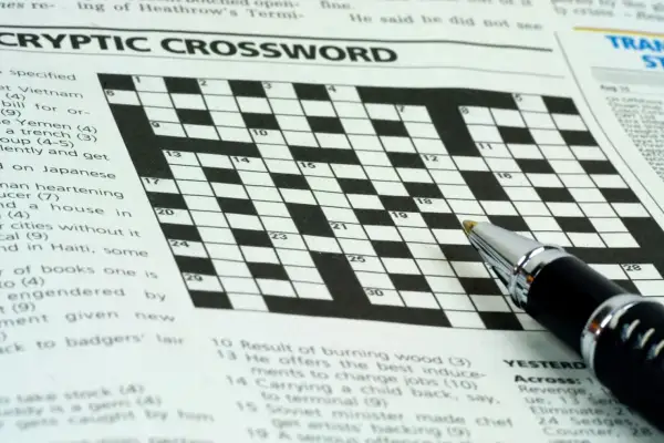 Zbliżenie na krzyżówkę z długopisem, widoczne nagłówki "CRYPTIC CROSSWORD" i fragmenty zadań.