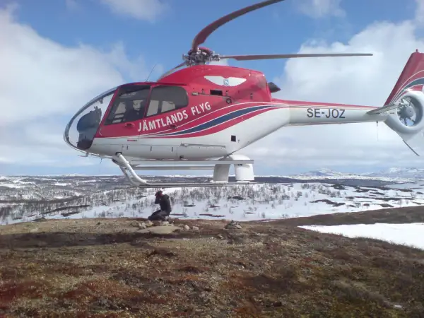Czerwony helikopter Jämtlands Flyg SE-JOZ wylądował na zaśnieżonym wzgórzu. Fotograf robi zdjęcie, uchwycony przez efekt rolling shutter.