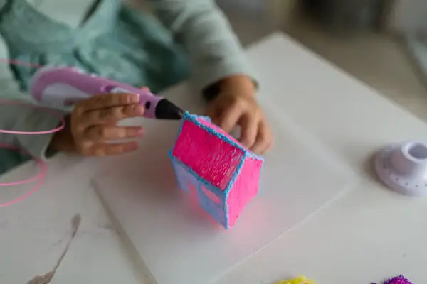 Dziecko tworzy różowy domek z niebieskim dachem za pomocą długopisu 3D. Powstaje piękny, trójwymiarowy ornament.