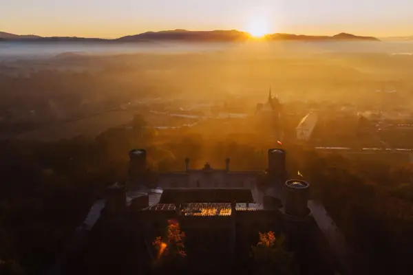 Zamek otoczony mgłą, skąpany w ciepłych promieniach **golden hour**. Słońce wschodzi nad wzgórzami, malując niebo na złoto.