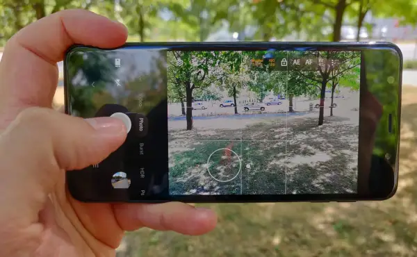 Dłoń trzyma smartfon z otwartą aplikacją do robienia zdjęć. Widok na drzewa i samochody.